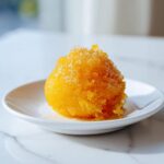 A bright orange scoop of Vegan Mango Pineapple Sorbet, topped with visible sugar crystals, served on a small white plate.