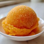 A vibrant scoop of bright orange Vegan Mango Pineapple Sorbet topped with coarse sugar crystals in a small white bowl.
