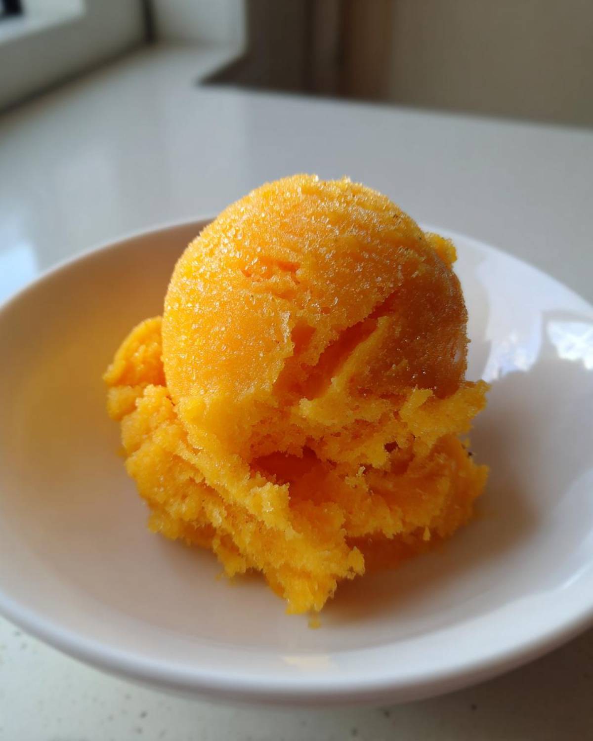 A bright orange scoop of icy Vegan Mango Pineapple Sorbet served in a small white bowl.