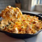 A wooden spoon lifts a serving of Cheesy Chicken and Rice Skillet, showing dramatic cheese pulls over the main dish in the cast iron pan.