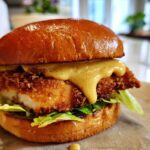 A close-up of a Crispy Chicken Burger with Honey Mustard Sauce dripping over the fried chicken patty and lettuce on a brioche bun.