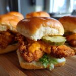 A close-up of several Crispy Chicken Sliders with Spicy Mayo, featuring a fried chicken patty, lettuce, pickle, and orange sauce on a soft bun.