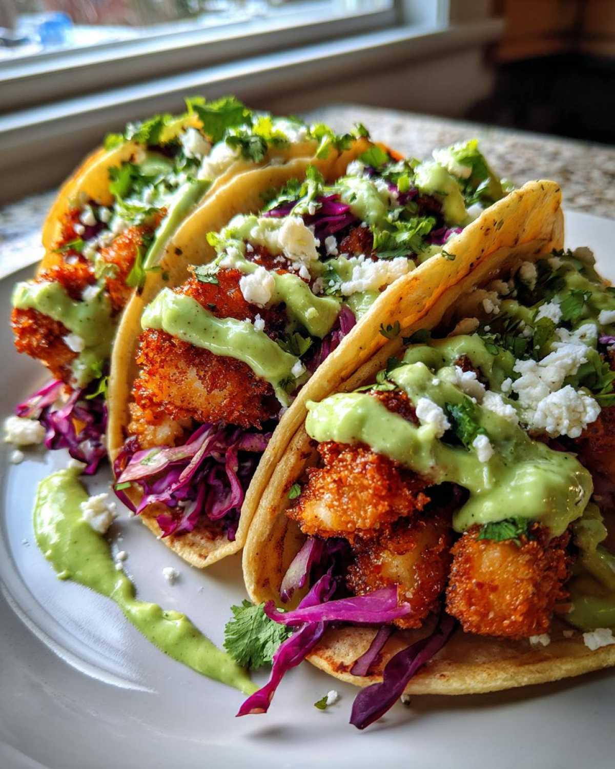 Three Crispy Chicken Tacos loaded with red cabbage, topped with avocado cream sauce and cotija cheese.