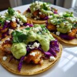 Four assembled Crispy Chicken Tacos topped with purple cabbage, avocado cream, and crumbled cheese on a white plate.