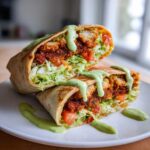 A Crispy Chicken Wrap with Avocado Sauce cut in half, showing crispy chicken, lettuce, tomatoes, and a generous drizzle of green sauce.