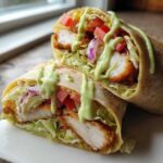 A halved Crispy Chicken Wrap with Creamy Avocado Sauce drizzled over crispy chicken, lettuce, tomatoes, and red onion.