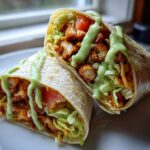 Two halves of a Crispy Chicken Wrap with Avocado Sauce, showing seasoned chicken, lettuce, and tomato drizzled with green sauce.