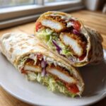 A halved Crispy Chicken Wrap showing crispy fried chicken, lettuce, tomato, red onion, and garlic yogurt sauce inside a tortilla.