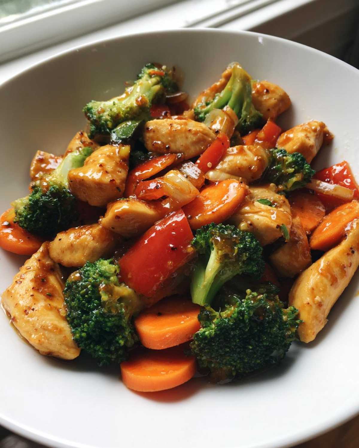 Close-up of a white bowl filled with Honey Garlic Chicken and Veggie Stir Fry featuring broccoli, carrots, and red peppers.