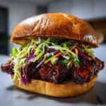 A close-up of a delicious Sticky BBQ Chicken Sandwich piled high with dark, glossy BBQ sauce and colorful crunchy slaw on a brioche bun.
