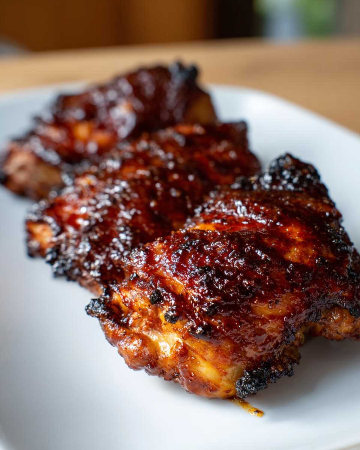 Three pieces of Sticky BBQ Chicken Thighs with crispy, caramelized edges resting on a white plate.