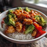 A close-up of a Sticky Orange Chicken Bowl with rice, bright green broccoli, and red bell peppers, topped with sesame seeds.