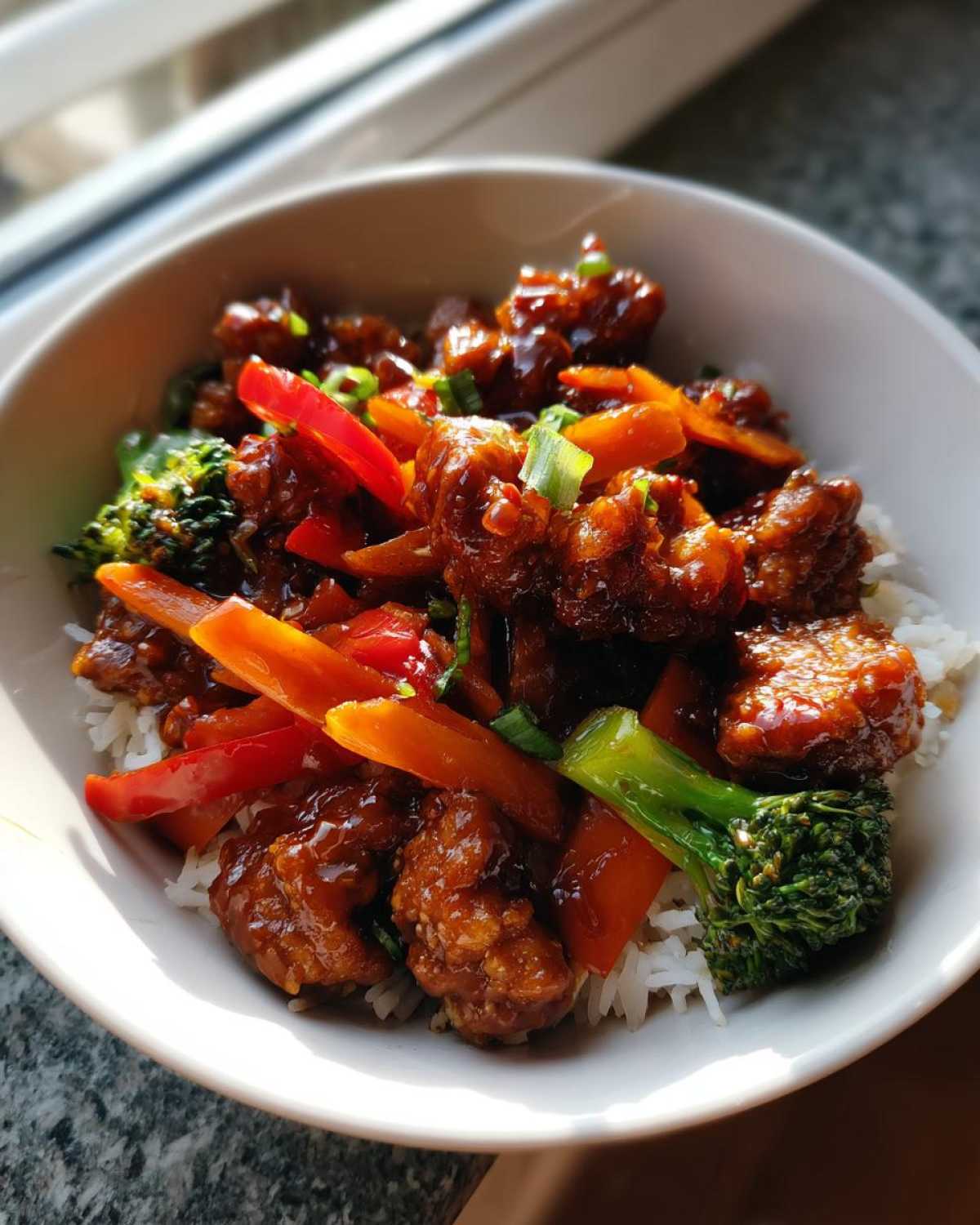 A close-up of a white bowl filled with Sticky Orange Chicken Bowl with Veggies served over white rice.