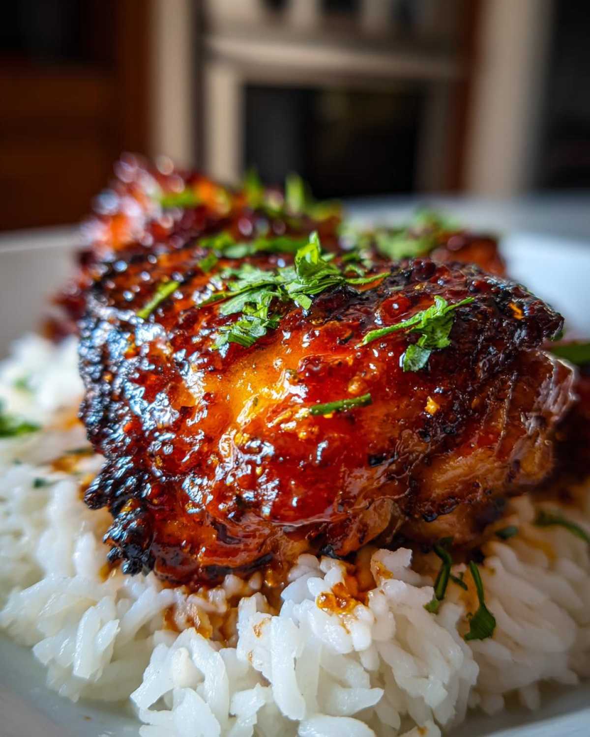 A close-up shot of a sticky sweet chili chicken thigh resting on a bed of white rice, garnished with fresh herbs.