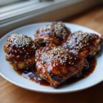 Four glazed Sticky Teriyaki Chicken Thighs with Garlic, generously topped with white sesame seeds on a white plate.
